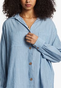 Light blue striped button-up shirt with a collar, two chest pockets, and wooden buttons. Fabric has a soft, textured appearance.