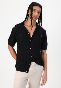 Schwarzes kurzärmliges Strickshirt mit Wellenmuster, mit einem flachen Kragen und Knöpfen. Tragen mit hellbeigen Leinenhosen.