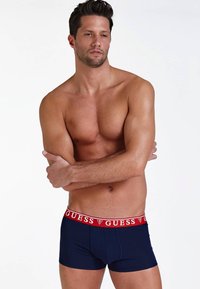 Boxeri scurți de culoare albastru marin, cu un elastic în talie pe care este înscris logo-ul roșu "GUESS". Materialul pare a fi neted și elastic, conceput pentru a oferi o siluetă ajustată.