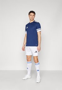 Maillot de sport bleu marine avec des manches rayées blanches, associé à un short blanc avec des accents noirs. Les accessoires comprennent des chaussettes blanches avec des rayures bleues.