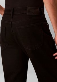 Άνδρας που φοράει μαύρα τζιν Calvin Klein, εστίαση στις πίσω τσέπες και την περιοχή της μέσης με ορατό διακριτικό της μάρκας.