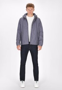 Grå mønstret jakke med hætte, i syntetisk materiale. Båret over en hvid t-shirt og parret med mørke jeans og hvide sneakers.