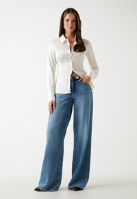 Camicia di raso bianca a maniche lunghe con chiusura a bottoni, abbinata a jeans in denim blu a gamba larga. Cintura nera con fibbia in argento.