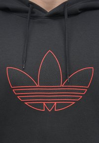 Svart hettegenser med en strukturert, mørk logo som har tre løvformer; konturert i lys rød med horisontale striper under.