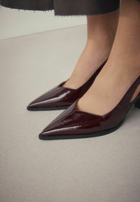 Burgundinės patentinės odos slingback kulniukai su smailiu priekiu, turintys išpjovos dizainą ir žemą blokuotą kulną, ant šviesiai pilkos paviršiaus.
