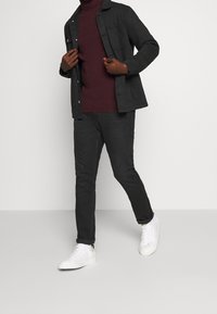 Schwarze Jacke über einem burgunderfarbenen Rollkragenpullover, kombiniert mit dunklen Hosen und weißen Sneakers. Die Materialien wirken leicht und haben eine glatte Textur.