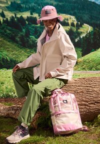 Lekka kremowa kurtka, zielone spodnie cargo, różowa kapelusz w kształcie wiadra i różowo-beżowy plecak z logo. Teksturowana tkanina, swobodny styl na świeżym powietrzu.