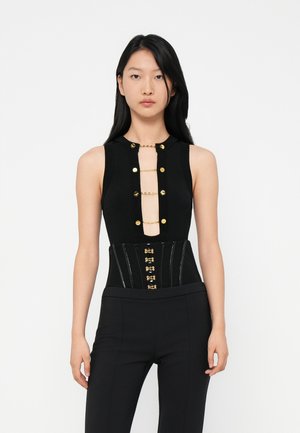 Young woman with long black hair wearing a sleeveless black top with gold chain details and black high-waisted pants against a white background.