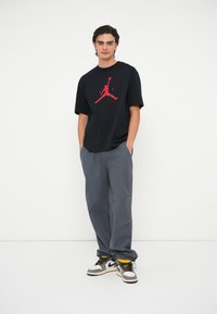 Must mustaheline T-särk mustade lühikeste varrukatega punase Air Jordani logoga, ühendatud avarate hallide pükstega ja mustade ja valgete sneakeritega, millel on kollased detailid.
