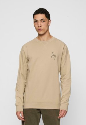 Beige long-sleeve sweatshirt with a crew neckline. Features a black hand graphic on the left chest. Soft fabric, relaxed fit, and ribbed cuffs.