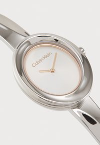 Montre argentée avec un cadran ovale, un cadran blanc, des aiguilles minimalistes en rose doré, et le logo "Calvin Klein". Bracelet en métal lisse.