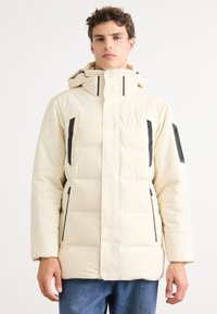 Beige Steppjacke mit einer Kapuze mit Reißverschluss, die schwarze Akzente und mehrere Taschen aufweist. Der Stoff wirkt glatt und isoliert.