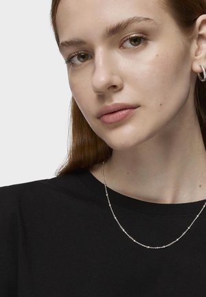 Collar de cadena de plata con pequeñas cuentas de metal, llevado sobre una blusa de tela negra. Textura suave y diseño minimalista.