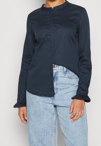 Blouse bleu marine à manches longues avec col rond, fermeture à boutons et poignets à froufrous, associée à un jean taille haute bleu clair. Texture de tissu lisse.