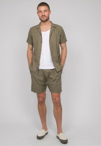 Chemise à manches courtes vert olive associée à un short assorti et un débardeur blanc. Chaussures décontractées à enfiler de couleur beige clair. Texture de tissu lisse.