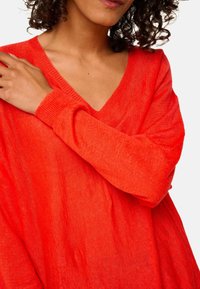 V-Ausschnitt, langärmeliger Pullover in einem lebhaften Orange, mit lockerer Passform und strukturiertem Strick. Das Material wirkt leicht und weich.