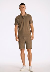 Brun piké med korta ärmar och krage, kombinerad med matchande shorts. Tyget har en strukturerad yta. Skorna är vita sneakers.