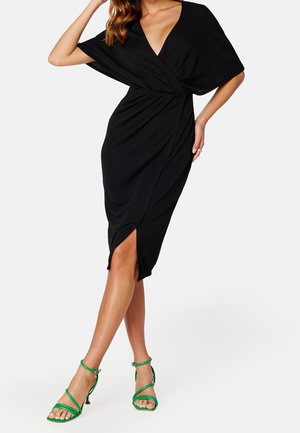 Femme portant une robe portefeuille noire avec un décolleté en V et des sandales vertes à talons hauts avec des brides, debout avec une jambe croisée devant l'autre.