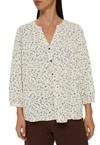 Femme portant une blouse crème avec un petit imprimé floral multicolore, boutonnée devant, manches trois-quarts, associée à un pantalon marron foncé.