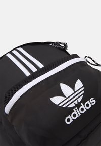 Černý batoh s bílými akcenty, včetně tří pruh ů a loga. Vyroben z texturované látky s detailem síťované kapsy a stahovací šňůrkou.