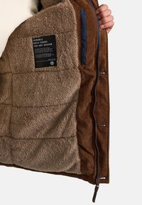 Brown corduroy jacket with a fuzzy beige lining, featuring a label, zip closure, and side pockets with blue accents and textured fabric.