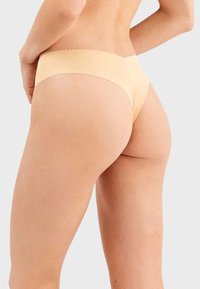 Tanga beige sin costuras, fabricado con una tela suave, que presenta una cintura con borde ondulado y un diseño de mínima cobertura. Textura suave con un ajuste que se adapta al cuerpo.