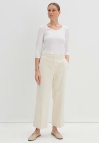 Weite, cremefarbene Cordhosen mit seitlichen Taschen und einem strukturierten Muster, kombiniert mit einem eng anliegenden weißen Langarmoberteil. Glatte flache Schuhe.