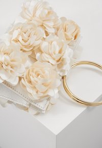 Petit sac à main blanc décoré de fleurs en tissu crème et d'une anse ronde en métal doré sur une surface blanche.