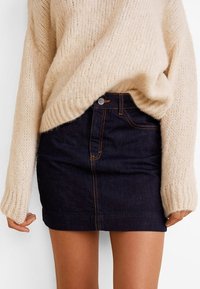 Jupe mini en denim bleu foncé avec des coutures orange, dotée d'une fermeture à boutons sur le devant et d'une coupe droite, associée à un pull en tricot beige.