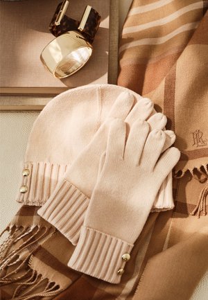 Gants en tricot beige et bonnet assorti avec des boutons dorés, une écharpe rayée marron à franges, et un bracelet manchette doré sur une surface en tissu.