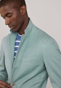 Blazer azzurro chiaro con colletto alla mandarina, chiusura a un bottone e taschino sul petto. Presenta una texture liscia. Sotto, è visibile una camicia a righe blu.