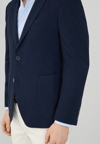 Blazer blu navy con una vestibilità su misura, due bottoni e una tasca. Presenta una texture liscia e un sottile dettaglio di cucitura sulla rever.