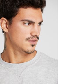 Sudadera gris claro con cuello redondo y tela texturizada; presenta costuras visibles y un primer plano de un vello facial y un tatuaje en el cuello.