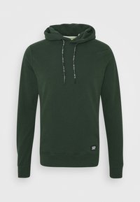 Sudadera de manga larga verde oscuro con un bolsillo frontal, cordones ajustables con texto blanco y un pequeño parche negro cerca del dobladillo.