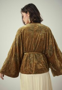 Brun velour kimono-stil jakke med gyldne perlebroderier af blomster, lange ærmer og et bundet bælte i taljen. Plisseret cremet nederdel synlig nedenunder.