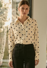 Chemisier à manches longues en crème avec des pois noirs, doté d'un col pointu, d'une fermeture à boutons et d'une coupe décontractée. Porté avec un pantalon noir taille haute.
