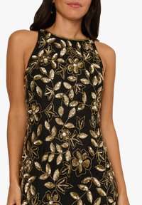Černé šaty bez rukávů s květinovými a listovými vzory z zlatých flitrů a krystalovými ozdobami na hrudi.