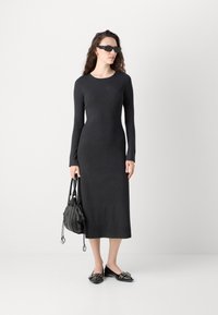 Long-sleeve, fitted black dress in a soft material, paired with a black bag and shoes featuring small decorative accents.