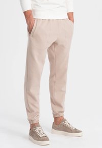 Beige Sweathosen aus einem weichen, strukturierten Material. Mit schrägen Seitentaschen, elastischen Bündchen und kontrastierenden Nähten. Pairing mit grauen Sneakers.