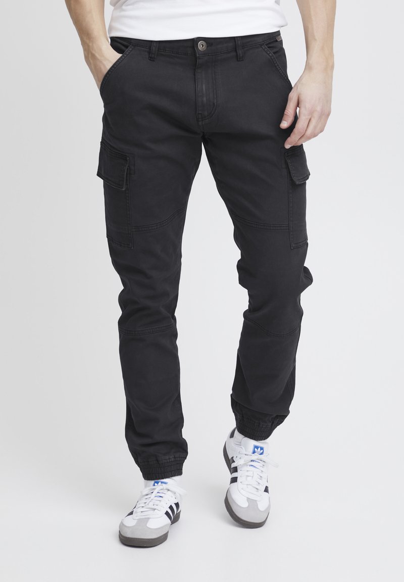 Sorte cargo-bukser med flere sidelommer, tætsiddende ankler, knaplukning og en letvægts, holdbar tekstur. Båret med sporty sneakers.