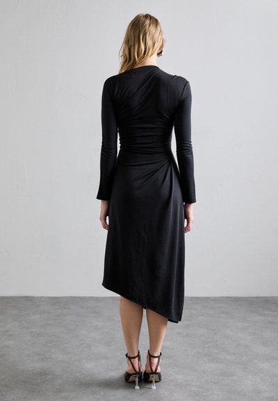 Vestido negro de manga larga con un cuerpo ajustado, detalles fruncidos y un dobladillo asimétrico. Tejido suave y elástico que se combina con sandalias de tacón alto.