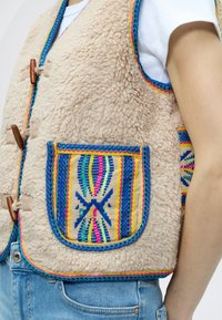 Γιλέκο από ψεύτικη γούνα σε μπεζ χρώμα με πολύχρωμες κεντητές λεπτομέρειες και τσέπη με σχέδιο. Διακοσμημένο με ξύλινα κουμπώματα και φωτεινές αντίθετες λεπτομέρειες.