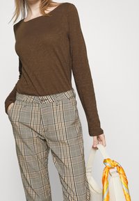 Pitkähihaisessa ruskeassa paidassa on neliönmuotoinen pääntie, joka on yhdistetty vaaleanruskeisiin ja ruudullisiin housuihin. Asusteina on vaaleanruskea laukku ja oranssi huivi.