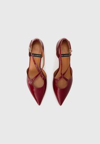 Chaussures en cuir verni rouge avec des orteils pointus, un design à sangle croisée et des côtés ouverts. Présentent une petite boucle sur la sangle de la cheville et une texture lisse.