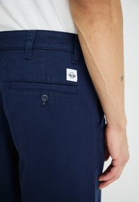 Navy cotton trousers with a flat back pocket and button closure. Features a brand label patch on the pocket. Smooth texture.