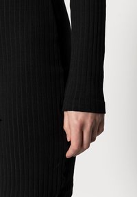 Svart ribbad långärmad plagg med en tät passform. Har en texturerad yta och en mudd i ärmslutet.