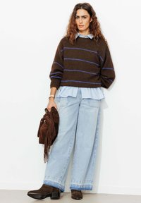 Pull en maille marron avec rayures bleues, superposé à une chemise bleu clair, associé à un jean denim clair à jambes larges et un sac à franges marron.