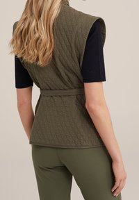 Tikattu oliivinvihreä liivi, jossa on korkea kaulus, hihaton malli ja vyötärönauha, yhdistettynä laivastonsiniseen ribbineulottuun lyhythihaiseen toppiin ja vihreisiin housuihin.