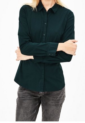 Person iført en mørkegrøn langærmet skjorte med knapper, med armene krydset over brystet og falmede sorte jeans.