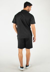 Polo-shirt noir à manches courtes, associé à un short noir. Modèle de dos, portant des chaussures blanches avec des accents noirs.
